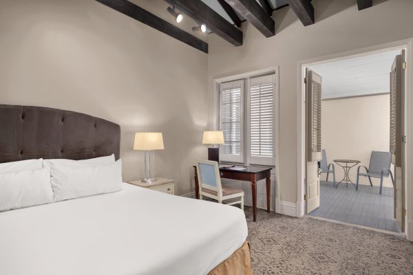 A cozy bedroom features a plush bed, desk, and lamps, leading to a balcony with chairs and a table, under wooden ceiling beams.
