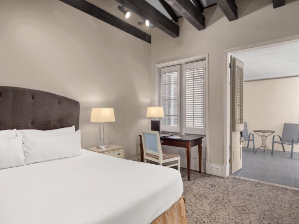 A cozy bedroom features a plush bed, desk, and lamps, leading to a balcony with chairs and a table, under wooden ceiling beams.