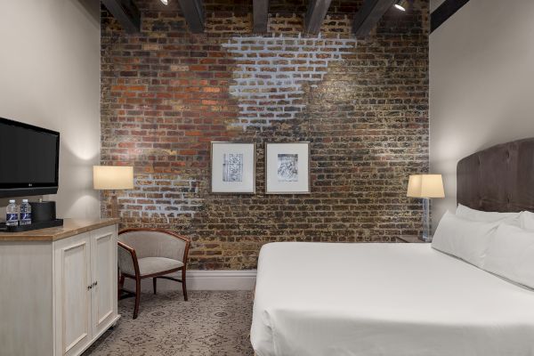 A cozy hotel room with a brick wall, bed, two lamps, and framed art; TV, water bottles, and a chair by a cabinet.