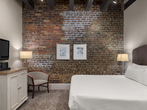 A cozy hotel room with a brick wall, bed, two lamps, and framed art; TV, water bottles, and a chair by a cabinet.