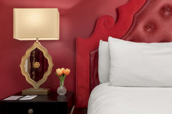 A red-themed bedroom scene with an ornate lamp, a small vase of tulips on a nightstand, and a white pillow on a bed.