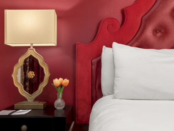 A red-themed bedroom scene with an ornate lamp, a small vase of tulips on a nightstand, and a white pillow on a bed.