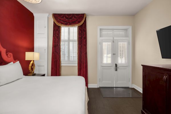 A hotel room with a white bed, red accents, a lamp, window with red curtains, door, and TV.