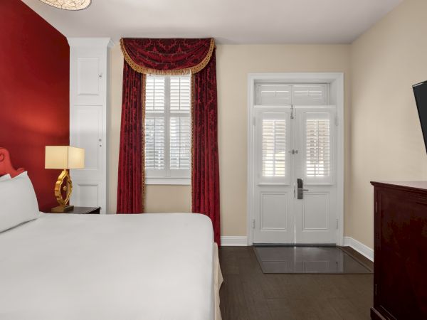 A hotel room with a white bed, red accents, a lamp, window with red curtains, door, and TV.