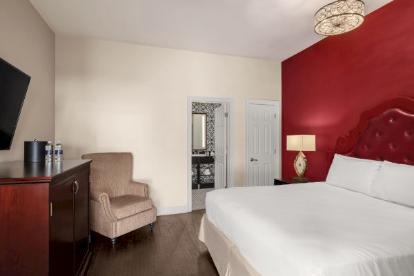 A hotel room with a red accent wall, bed, armchair, TV, and bathroom visible through an open door, with modern decor.