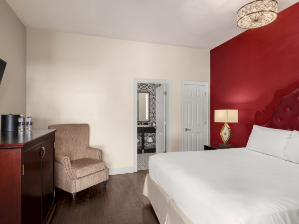 A hotel room with a red accent wall, bed, armchair, TV, and bathroom visible through an open door, with modern decor.