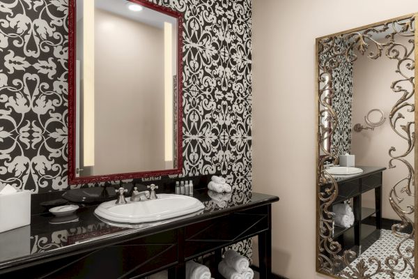 Ornate bathroom with patterned wallpaper, large mirrors, a sink, towels, and toiletries on a black countertop.