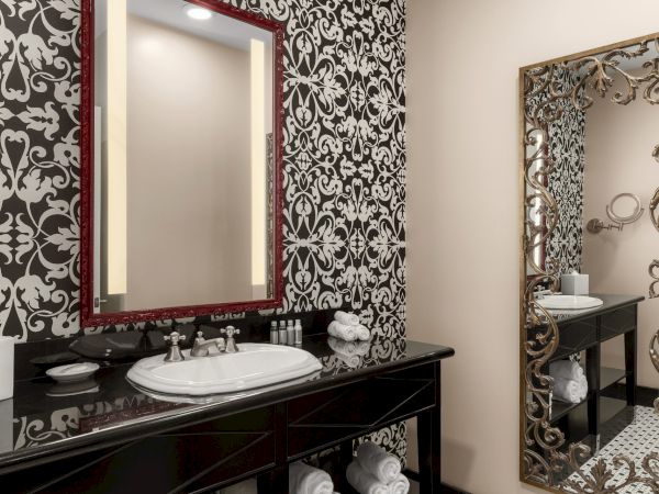 Ornate bathroom with patterned wallpaper, large mirrors, a sink, towels, and toiletries on a black countertop.