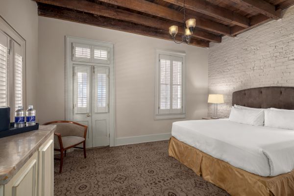 A cozy room features a bed, wooden ceiling, exposed brick wall, chair, lamps, and bottled water on a counter.