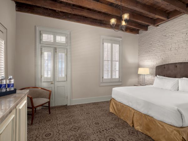 A cozy room features a bed, wooden ceiling, exposed brick wall, chair, lamps, and bottled water on a counter.