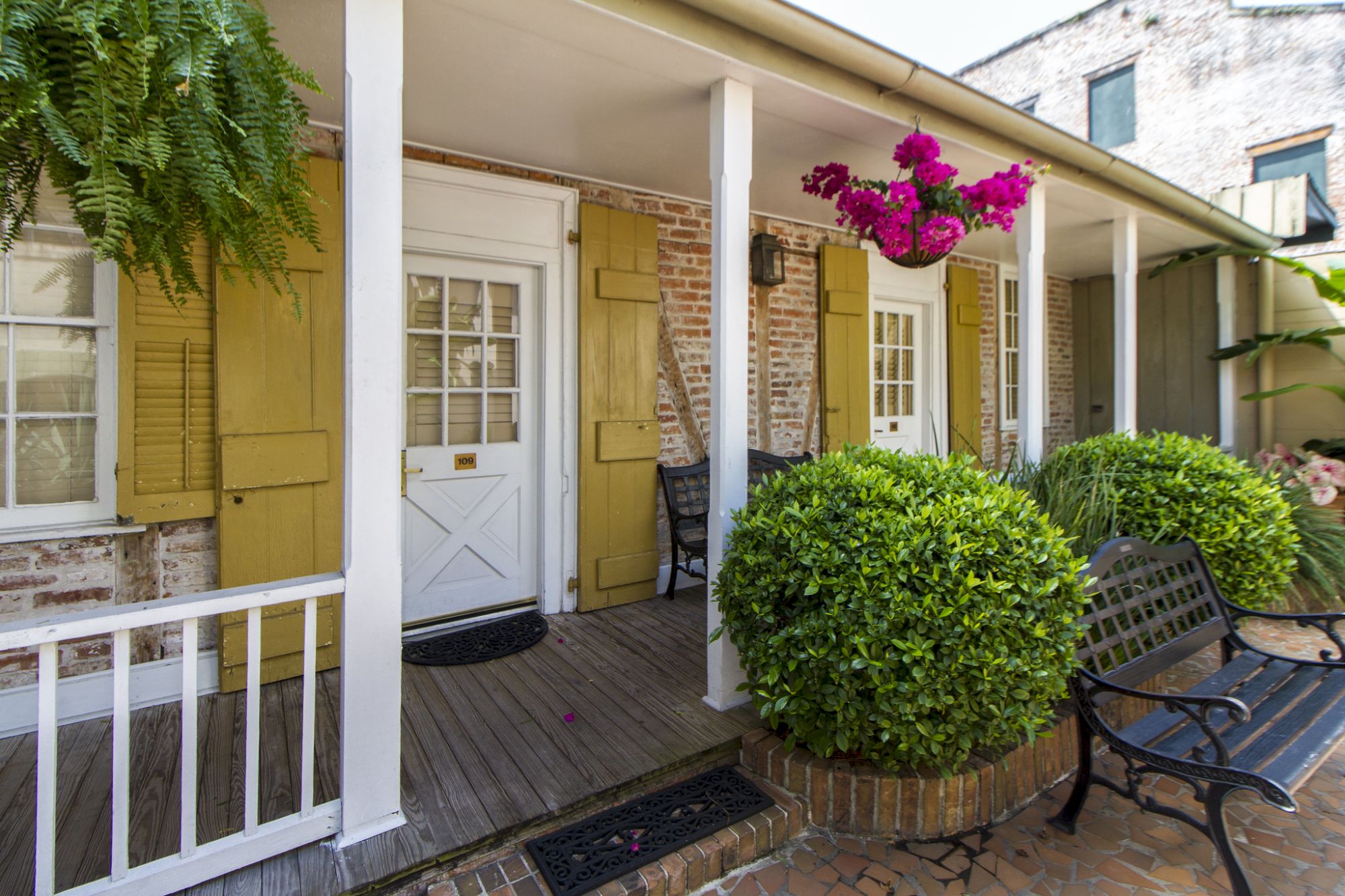 A charming porch with white doors, green shutters, hanging plants, and benches on a brick path.