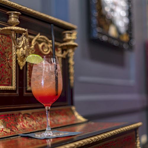 A cocktail with a lime garnish sits on an ornate piano, featuring a reddish and gold design, with a blurred background.