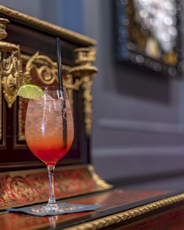 A cocktail with a lime garnish sits on an ornate piano, featuring a reddish and gold design, with a blurred background.