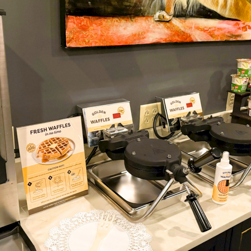 Waffle makers, a drink dispenser, assorted cups, a bowl with items, and a decorative painting are on a countertop.