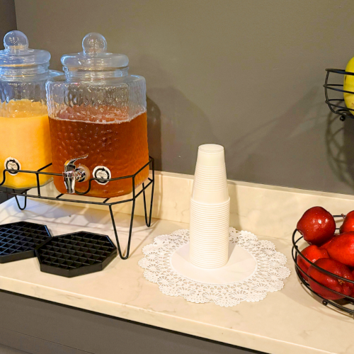 Two beverage dispensers, one with orange juice and the other with tea, are next to stacked cups. Baskets hold bananas and apples.