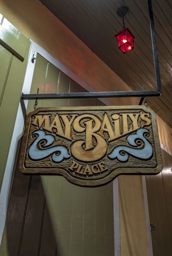 A hanging sign reads "May Baily's Place" with decorative design elements. A red light is above, against a wooden facade.