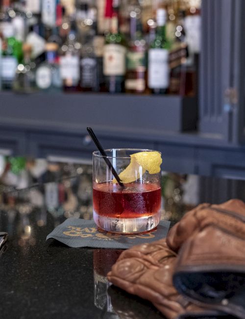 A cocktail with a lemon garnish and straw sits on a bar counter, accompanied by gloves and a wallet, with bottles in the background.