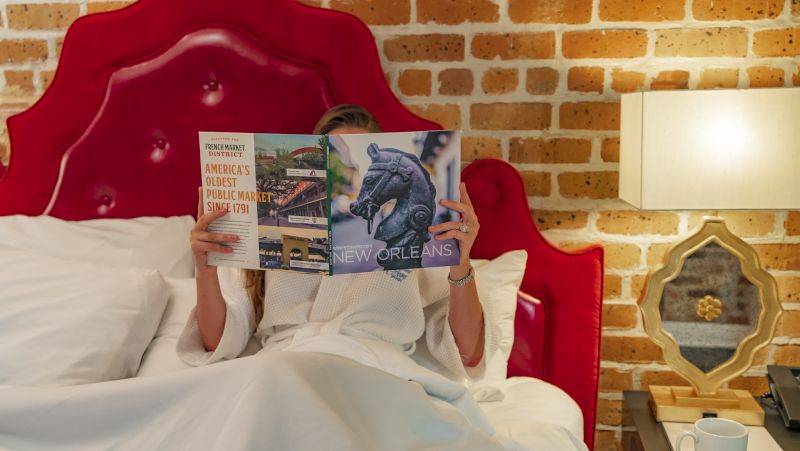 A woman sits in bed with a magazine in front of her face, in a room with a red leather headboard and brick wall.