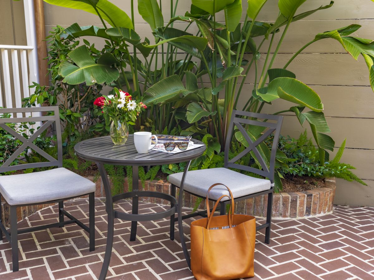 A patio scene with two chairs, a table, flowers, a mug, sunglasses, a magazine, and a tote bag, surrounded by lush green plants.