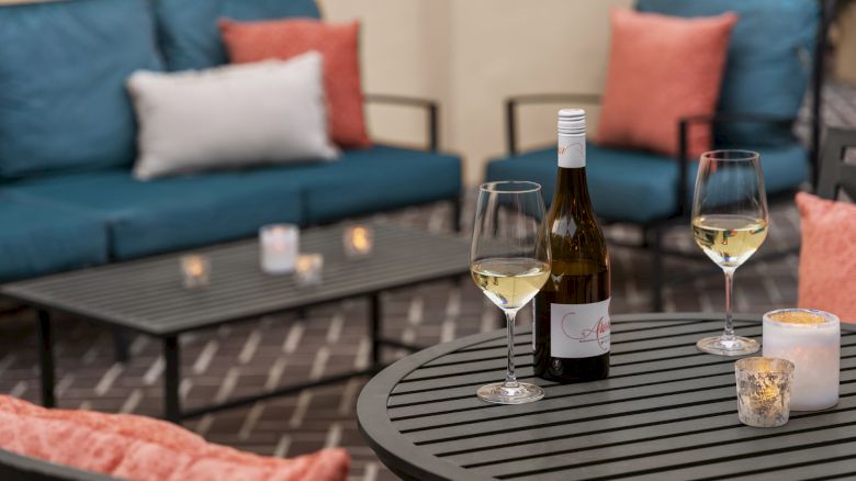 A cozy patio setting with a bottle of white wine, two glasses on a table, candles, and blue and orange cushions.
