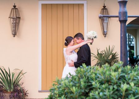 A couple in wedding attire embraces in front of a beige building with yellow shutters, surrounded by plants and lanterns.