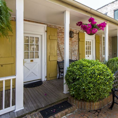 A cozy porch with two white doors, yellow shutters, potted plants, a hanging basket, and a black bench on brick flooring.