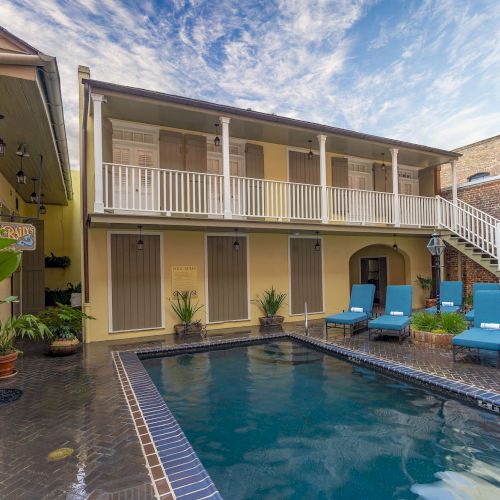 A courtyard with a pool, lounge chairs, plants, and a two-story building with a balcony on a clear day.