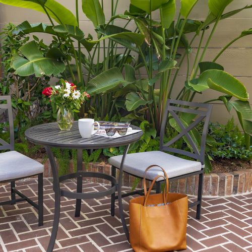 A patio with two chairs, small table with flowers, mug, sunglasses, magazines; surrounded by plants, and a tan bag on the ground.
