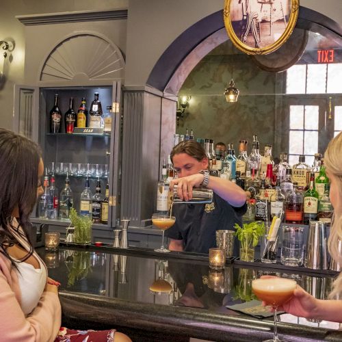 A bartender prepares drinks for two women at a well-stocked bar with elegant decor and a cozy atmosphere.