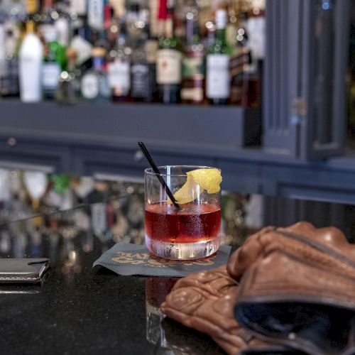 A cocktail with lemon garnish on a bar, accompanied by gloves and a wallet, with a selection of bottles in the background.
