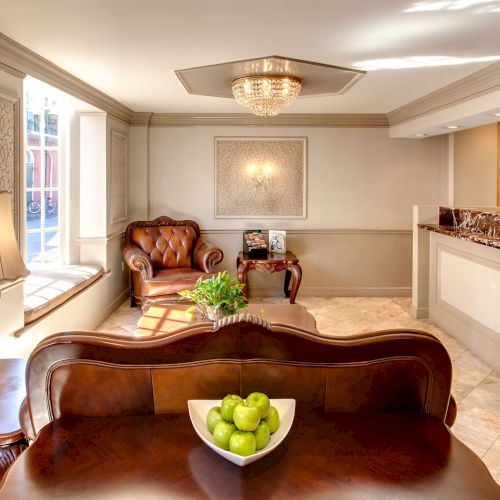 The image shows a cozy reception area with ornate furniture, a chandelier, a bowl of green apples, and a guest services desk.