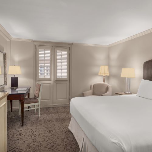 A hotel room with a bed, desk, TV, two lamps, and a soft chair; neutral tones throughout.