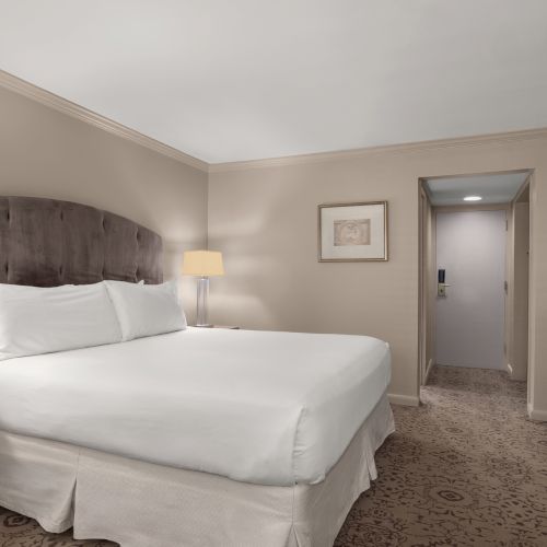 The image shows a hotel room with a large bed, nightstands, lamps, a dresser, and a hallway leading to a door.