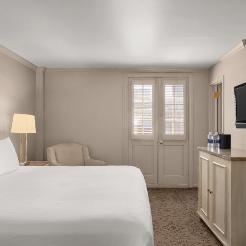 A tidy hotel room with a bed, lamp, armchair, TV, and a cabinet near double doors. The decor is neutral and minimalistic.