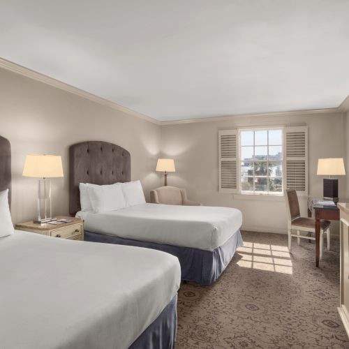 The image shows a hotel room with two beds, lamps, a desk with a chair, a TV on a cabinet, and a window with shutters.