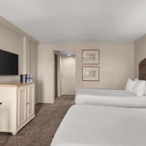 The image shows a hotel room with two beds, a TV on a cabinet, a desk, lamp, and framed pictures on a wall.