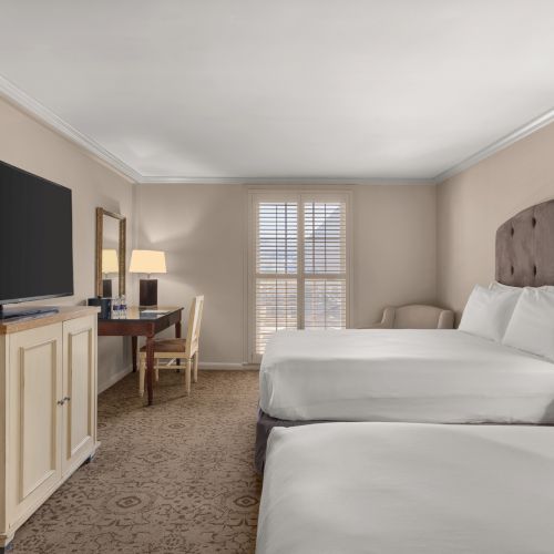 A hotel room with two beds, a large TV on a cabinet, a desk with a chair, lamps, and a window with shutters at the end of the room.