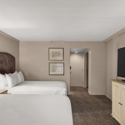 The image shows a hotel room with two double beds, a large TV on a cabinet, paintings on the wall, and a lamp on a nightstand.