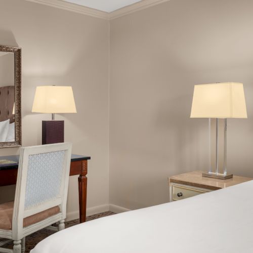 A hotel room with a bed, nightstand, lamp, desk, chair, mirror, and bottled water on the desk.