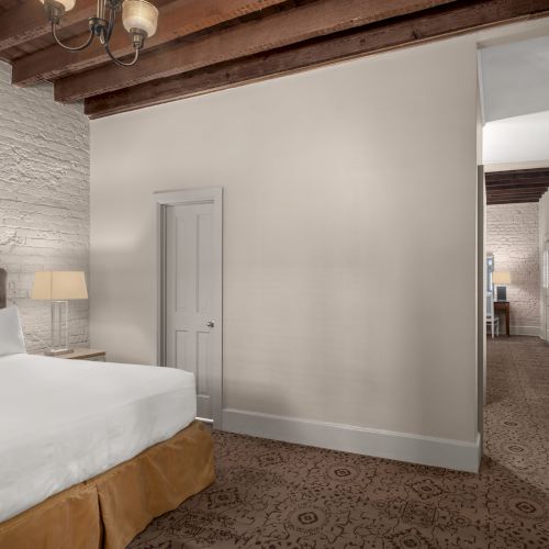 A cozy bedroom with a large bed, white brick wall, wooden ceiling beams, and a hallway leading to a lit area.