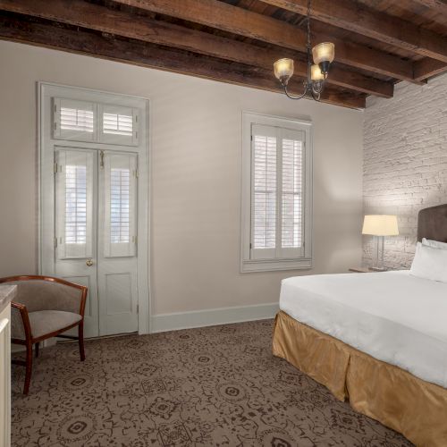 A cozy hotel room with a bed, wooden beamed ceiling, brick wall, chair, lamp, and a door with shutters.