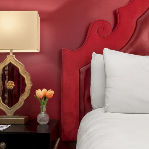 A bed with white pillows, a red headboard, a nightstand with a lamp, a vase with orange flowers, and a wall painted red.
