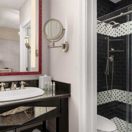 The image shows a bathroom with a vanity, sink, mirror, toiletries, and a glass-enclosed shower with black and white tile accents.