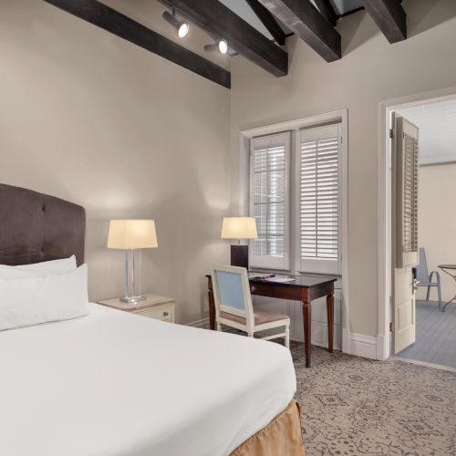 The image shows a neatly arranged bedroom with a bed, side table, lamp, desk, and chairs in an adjoining room.