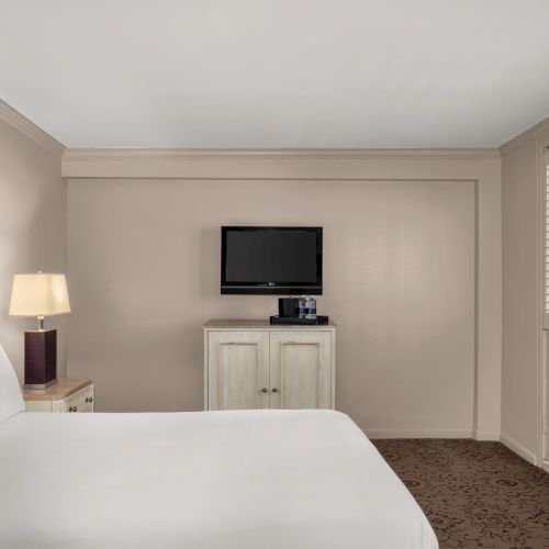 A hotel room with a bed, nightstand, lamp, wall-mounted TV, dresser, and window with shutters is shown. The decor is simple and neutral.