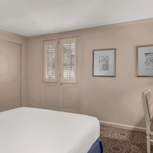 A hotel room features a bed, TV, desk, and chairs. There are framed pictures on the wall, and a window with shutters behind.