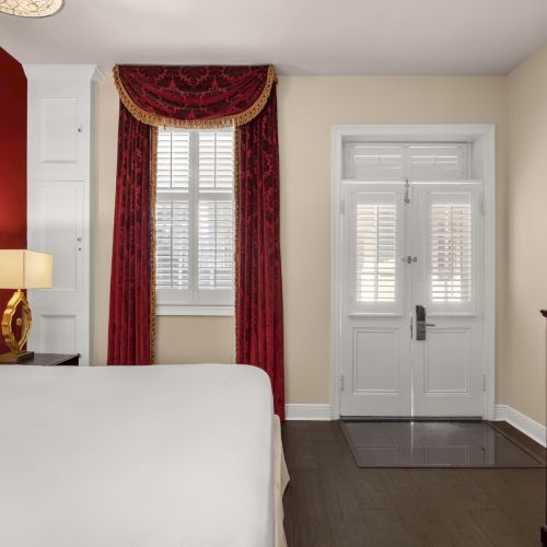 A hotel room with a red and white color scheme, featuring a bed, curtains, a lamp, and a flat-screen TV on a dresser.