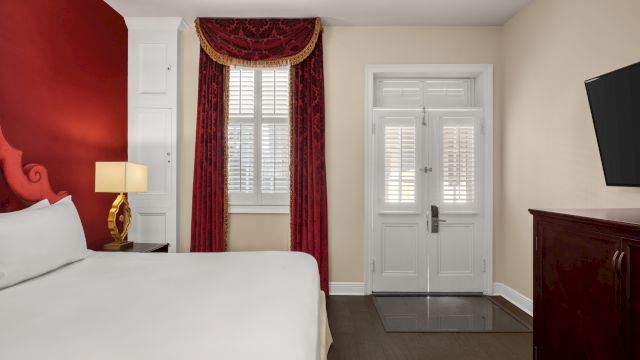 A hotel room with a white bed, a window with red curtains, a door, and a TV on a dresser.
