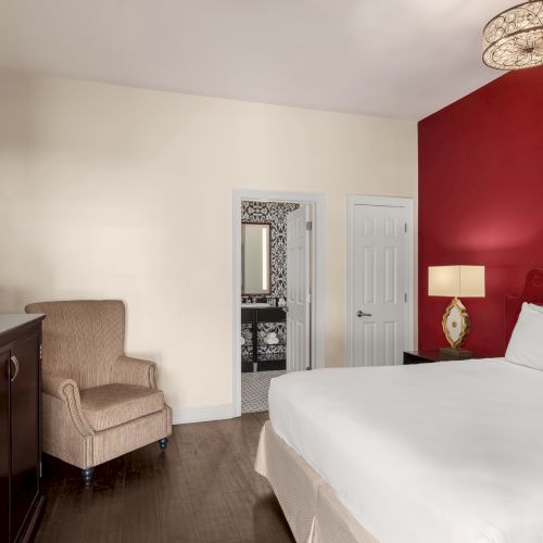 A hotel room with a bed, armchair, wall-mounted TV, red accent wall, lamp, and open door leading to a bathroom.