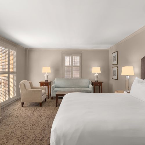 A cozy hotel room with a large bed, armchair, TV, sofa, and two lamps near a window with shutters enhances the ambiance.